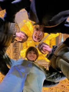 Family of four taking a selfie from below, framed by the lit ironwork of the Eiffel Tower at night